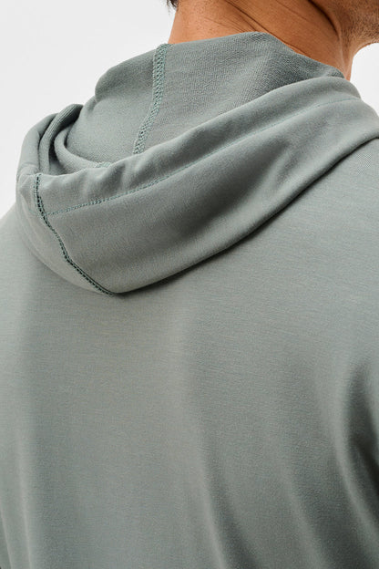 Close-up back view of a person wearing a sage green hooded sweatshirt, showing the hood, shoulder seam, and neck against a plain light background.