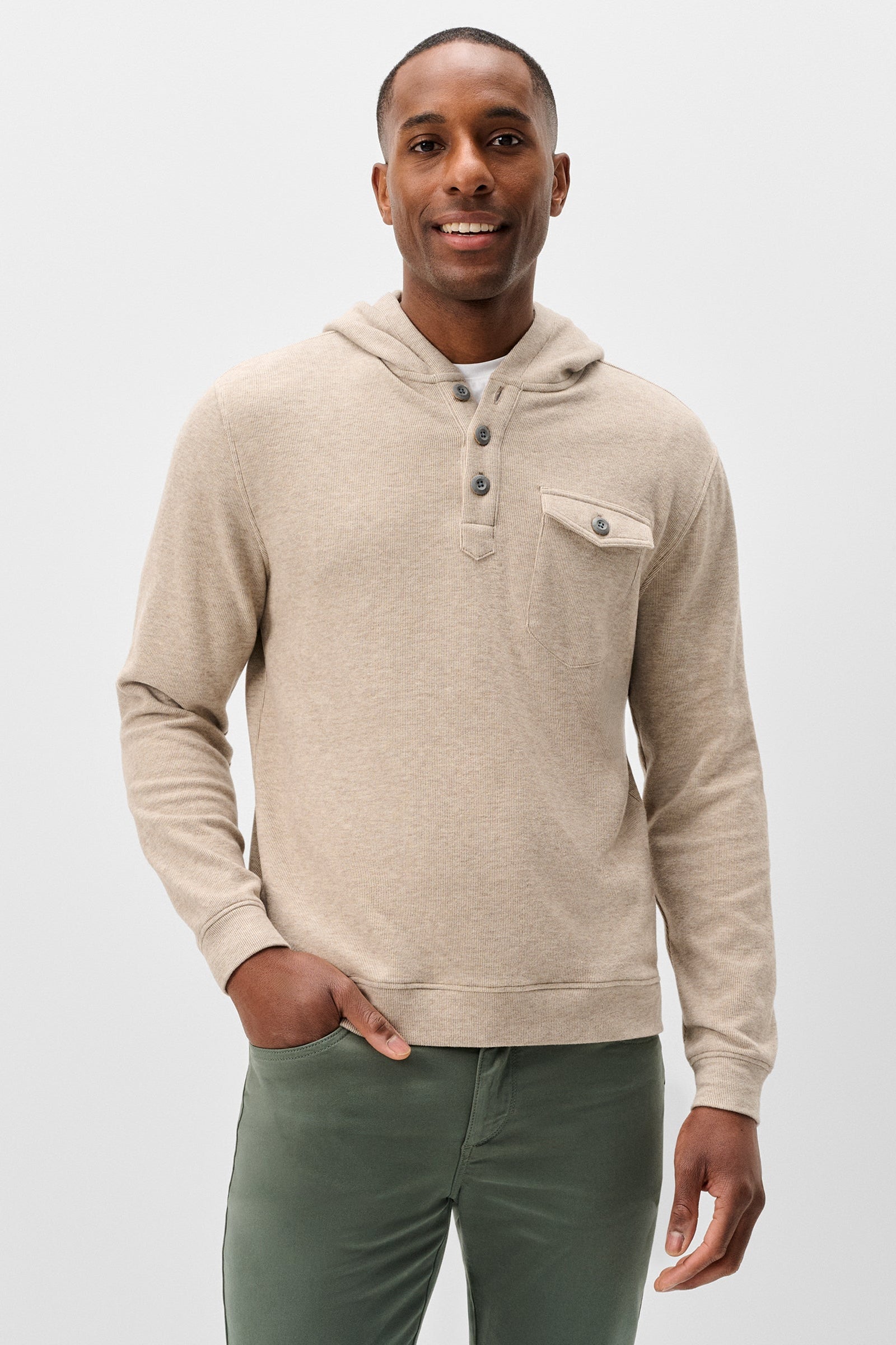 A man wearing a beige hooded knit sweater with a three-button henley collar and a chest pocket, paired with olive green pants, against a light gray studio background.