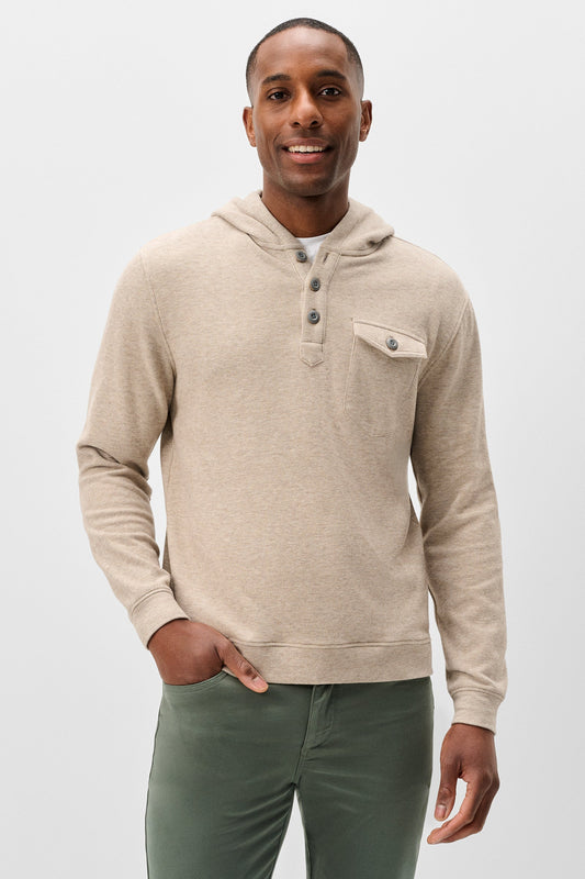 A man wearing a beige hooded knit sweater with a three-button henley collar and a chest pocket, paired with olive green pants, against a light gray studio background.