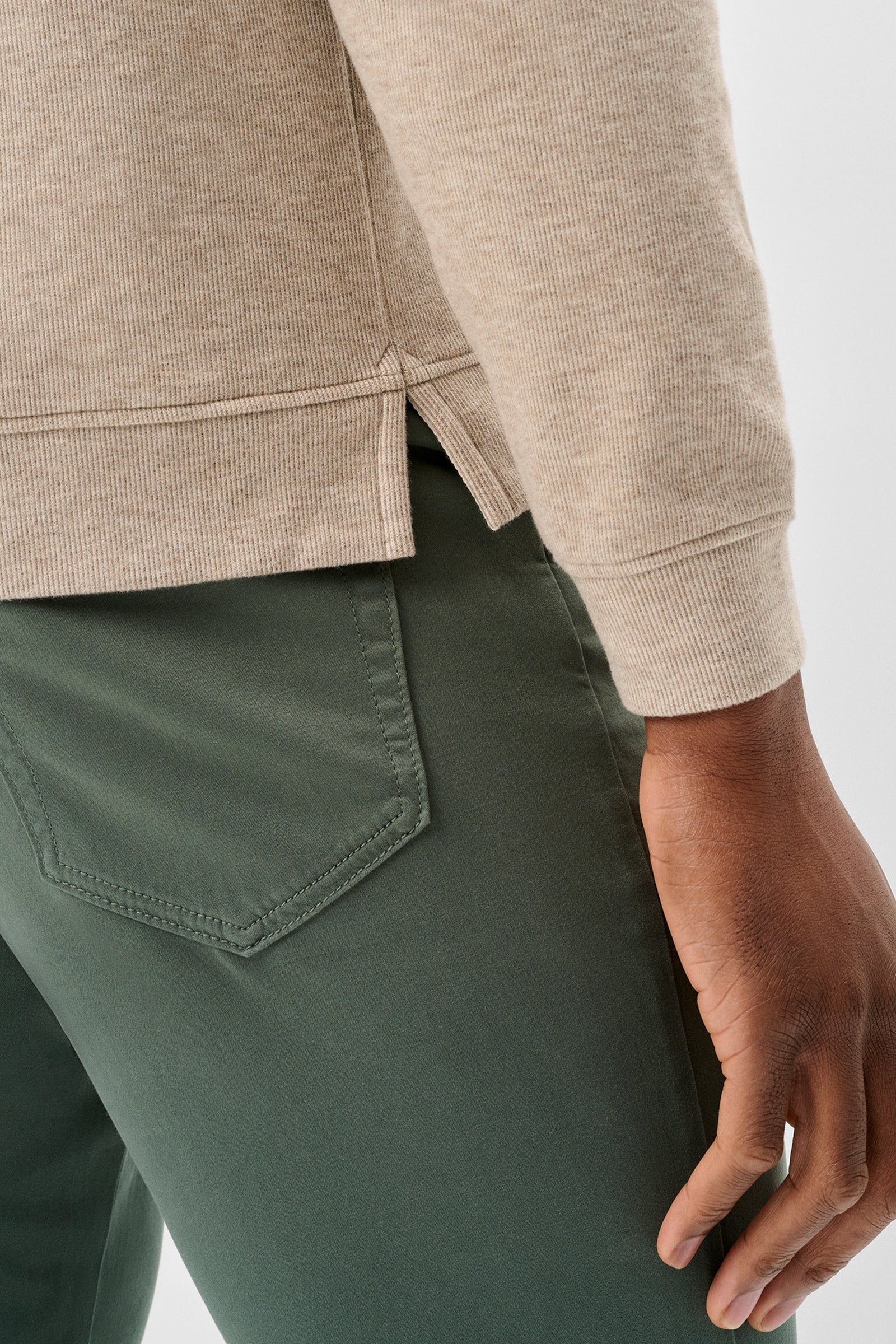 Close-up of a person wearing a light beige knit top and dark green pants, showing the side seam detail and back pocket of the trousers against a light gray background.