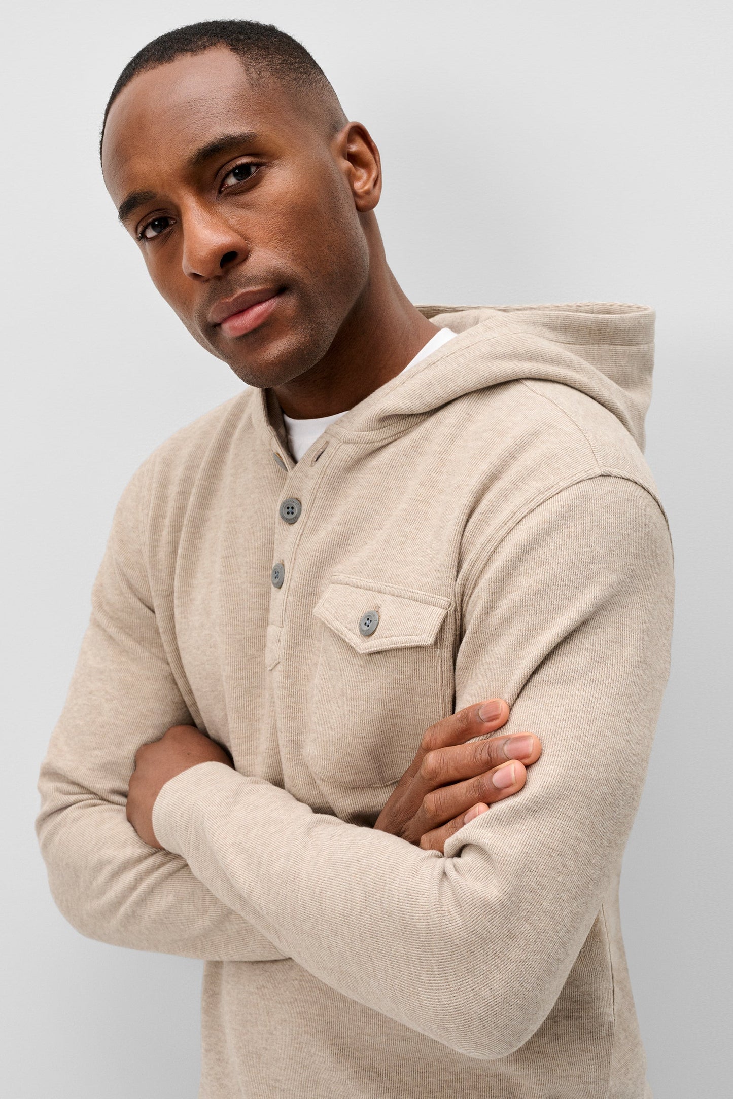 A man wearing a light beige hooded long-sleeve henley shirt with a chest pocket and button details, posed against a neutral light gray background.
