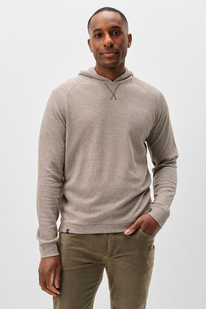 A man wearing a light taupe heathered pullover hoodie with raglan sleeves and olive green pants, standing against a plain light gray studio background.