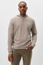 A man wearing a light taupe heathered pullover hoodie with raglan sleeves and olive green pants, standing against a plain light gray studio background.
