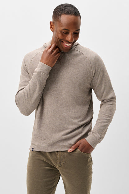 A man wearing a light taupe heathered long-sleeve pullover sweater and olive green pants, standing against a light gray studio background.