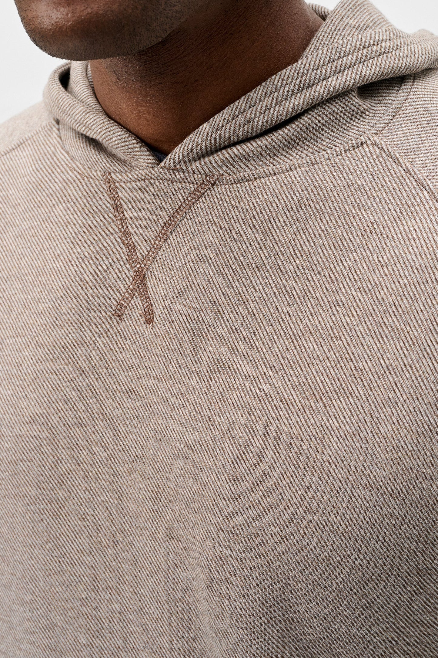 Close-up of a person wearing a textured beige hooded sweatshirt with a tonal embroidered X logo on the chest, set against a plain light gray background.