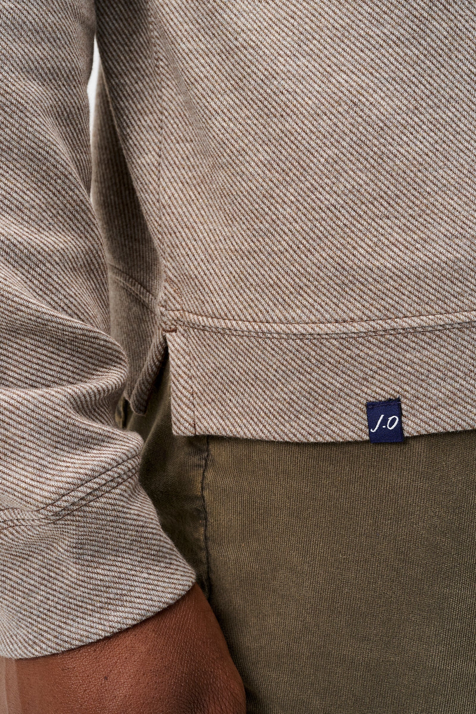 Close-up of a person wearing a textured taupe-beige long-sleeve top with a small navy blue 'J.O' brand tag near the hem, paired with olive green pants.