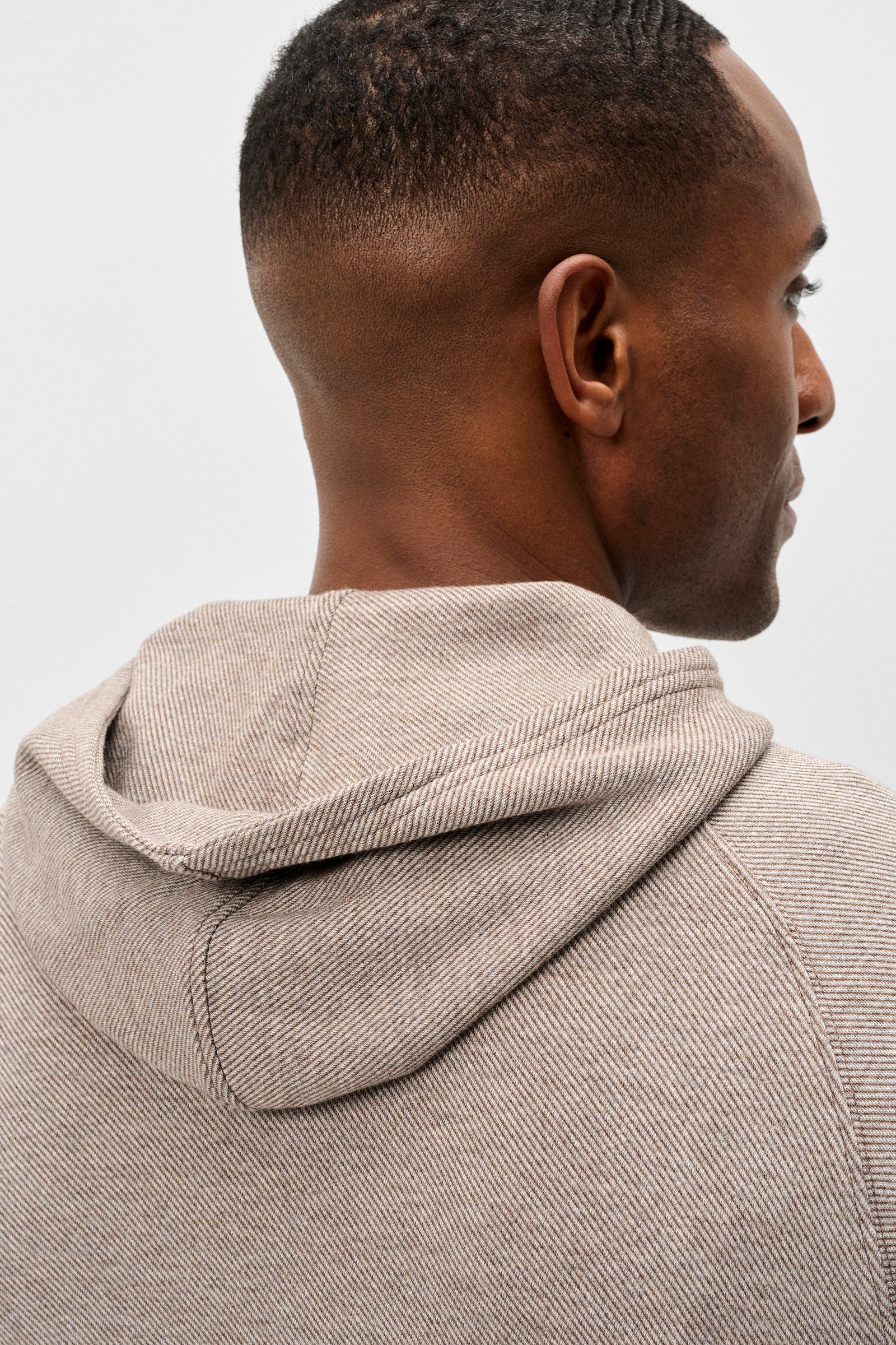 Close-up back and side view of a man wearing a light beige textured hooded sweatshirt against a plain light gray background.