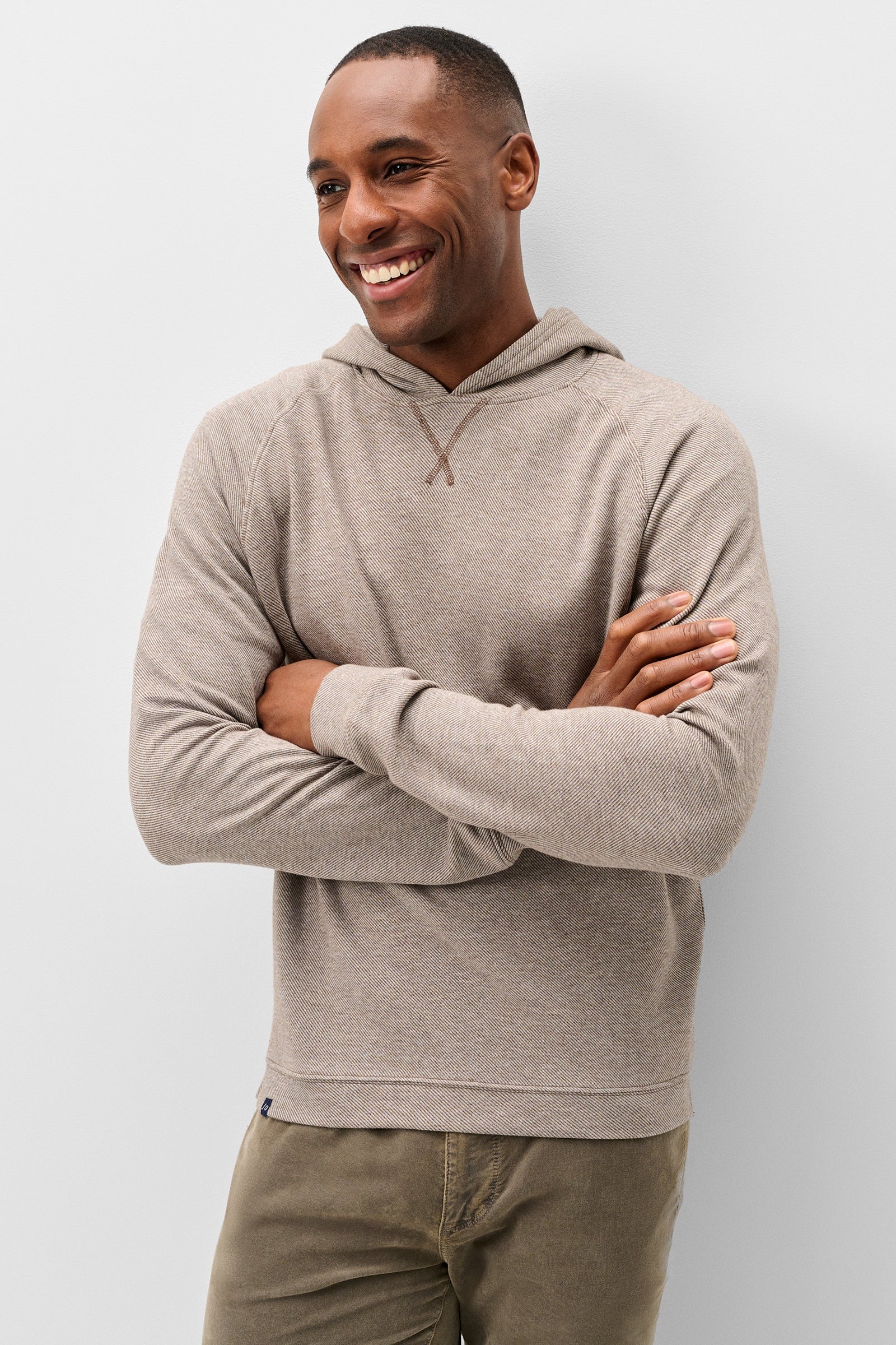 A smiling man wearing a light taupe heathered long-sleeve hoodie with a drawstring detail at the neck, paired with olive green pants, standing against a light gray studio background.