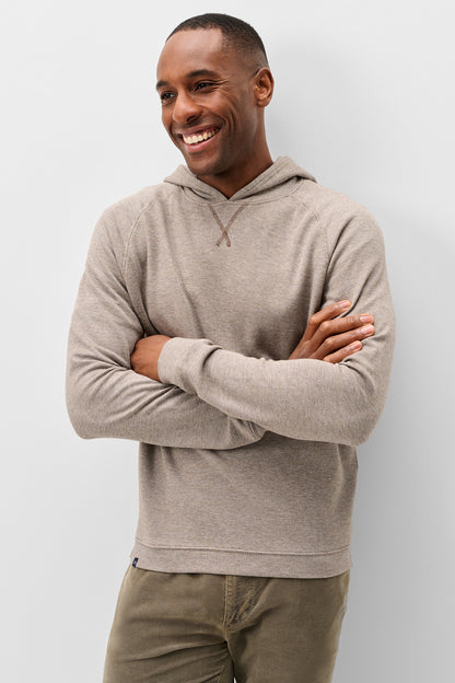 A smiling man wearing a light taupe heathered long-sleeve hoodie with a drawstring detail at the neck, paired with olive green pants, standing against a light gray studio background.