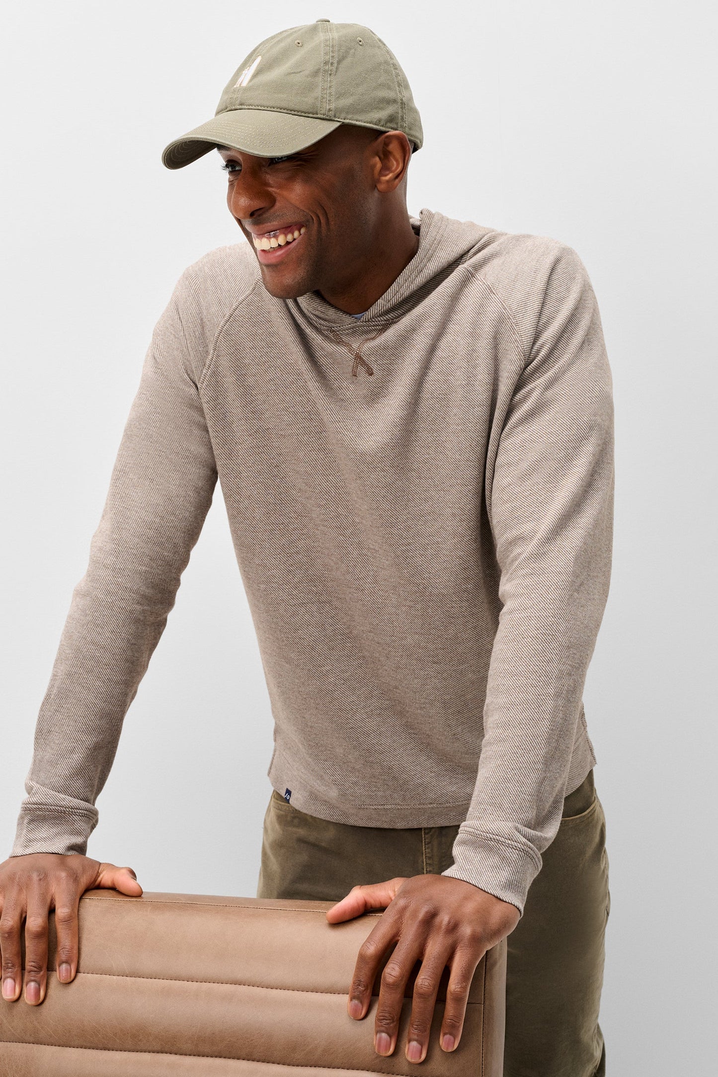 A man wearing a light taupe long-sleeve hooded shirt and an olive green baseball cap, smiling while leaning on a matching beige chair against a light gray studio background.