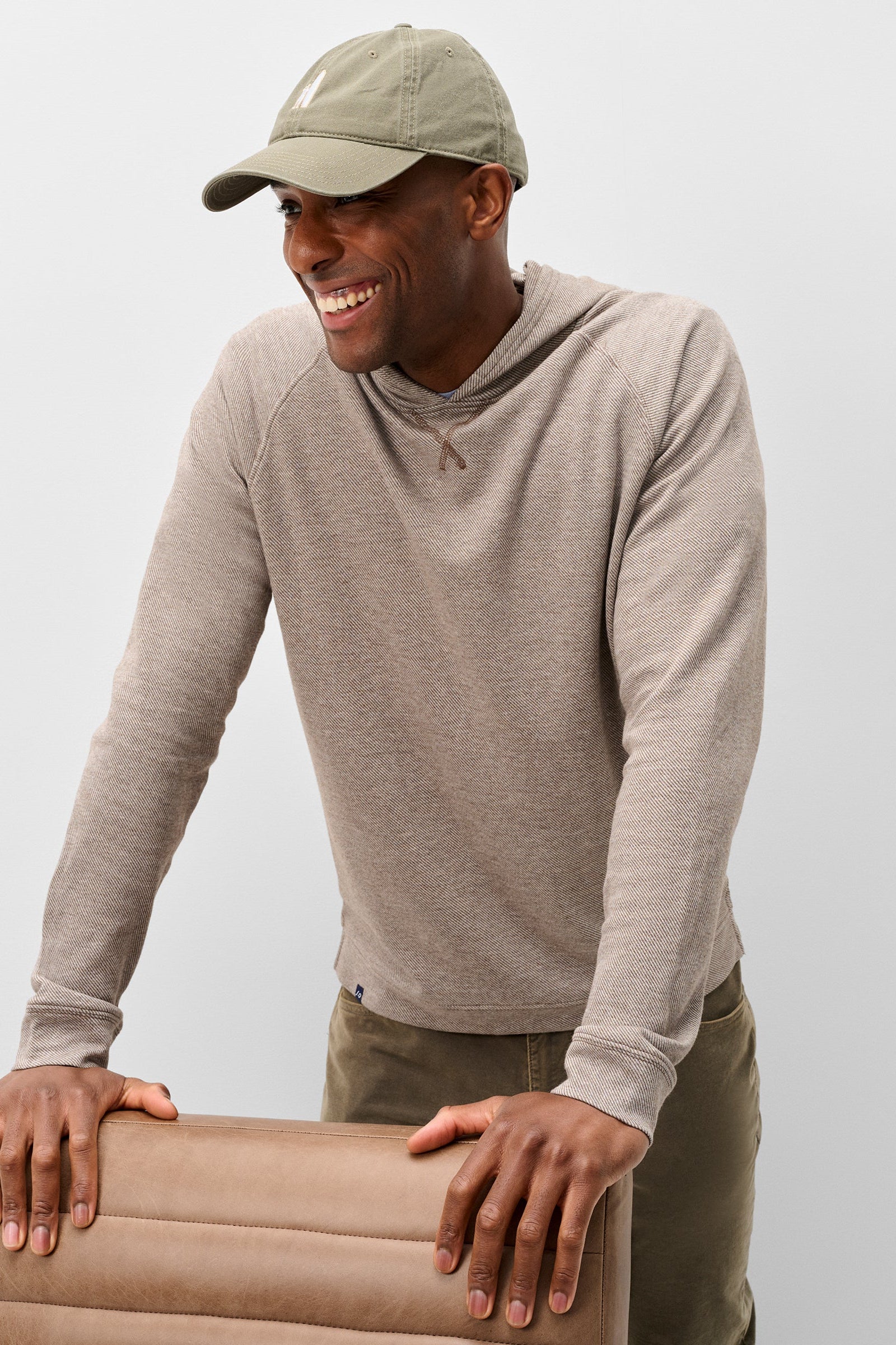 A man wearing a light taupe long-sleeve hooded shirt and an olive green baseball cap, smiling while leaning on a matching beige chair against a light gray studio background.