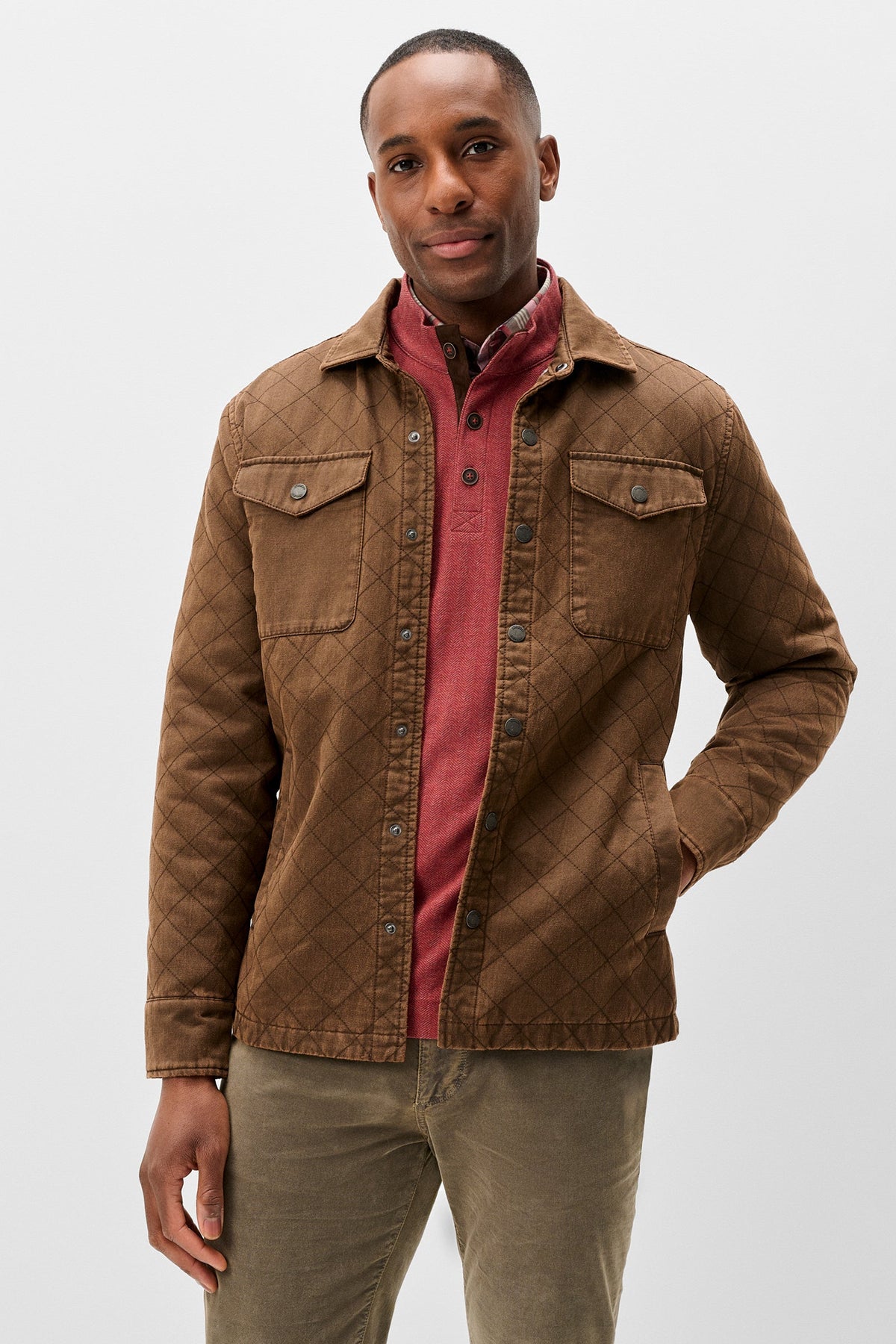 A man wearing a brown quilted overshirt jacket over a dusty red collared shirt and olive green trousers, standing against a light gray studio background.