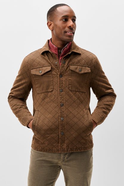 A man wearing a brown diamond-quilted corduroy jacket over a red collared shirt and olive pants, standing against a light gray studio background.