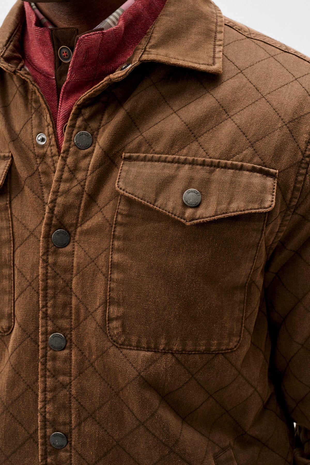 Close-up of a brown quilted overshirt with a diamond pattern, featuring a button-down front, chest pockets, and a red collared shirt underneath.