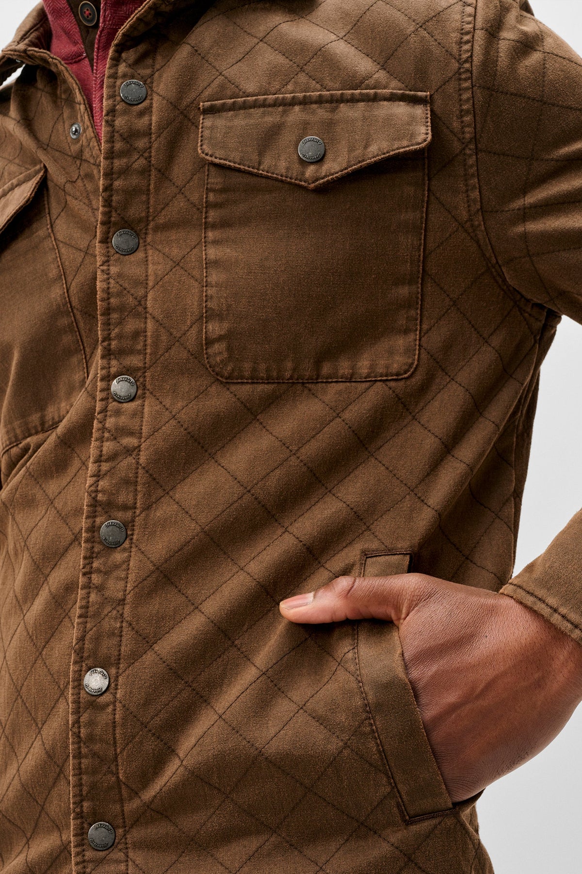 A close-up of a brown quilted button-up shirt jacket with a diamond pattern, featuring chest pockets and a hand tucked into a side pocket, set against a light gray background.