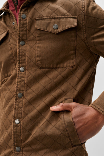 A close-up of a brown quilted button-up shirt jacket with a diamond pattern, featuring chest pockets and a hand tucked into a side pocket, set against a light gray background.