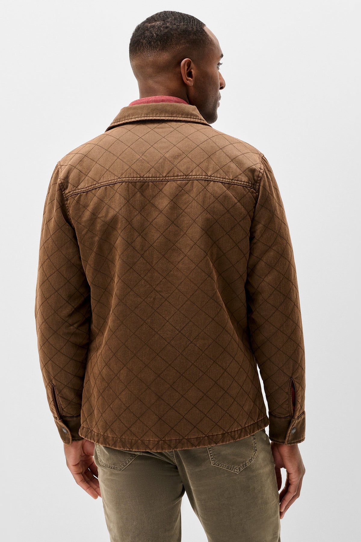 A man viewed from the back wearing a brown diamond-quilted jacket over a pink shirt and olive green pants, against a light gray studio background.