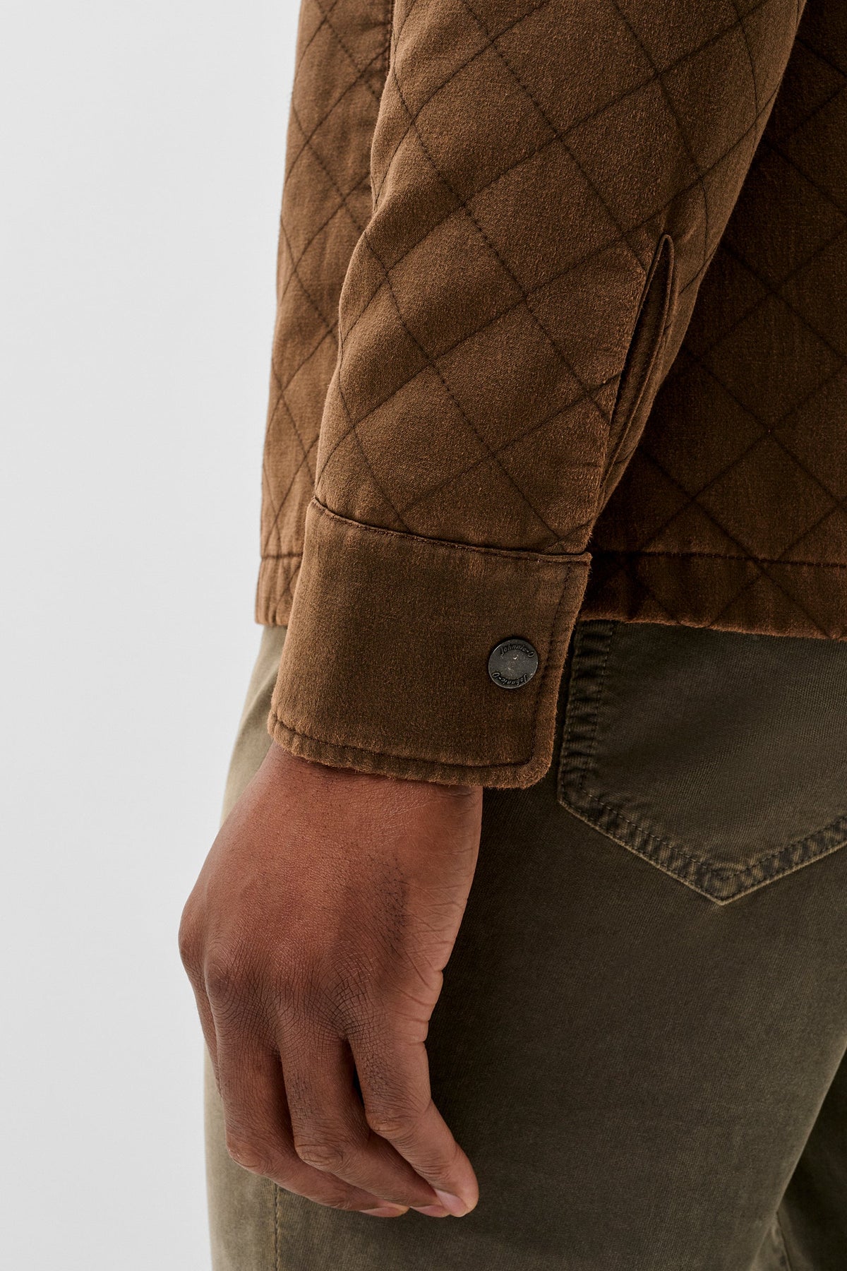 Close-up of a person wearing a brown quilted jacket with a buttoned cuff and olive green pants against a light gray background.