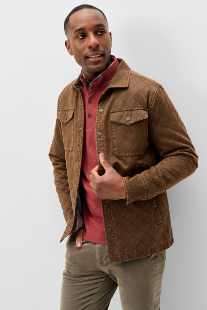 A man wearing a brown quilted corduroy jacket over a red shirt and olive pants, standing against a light gray studio background.
