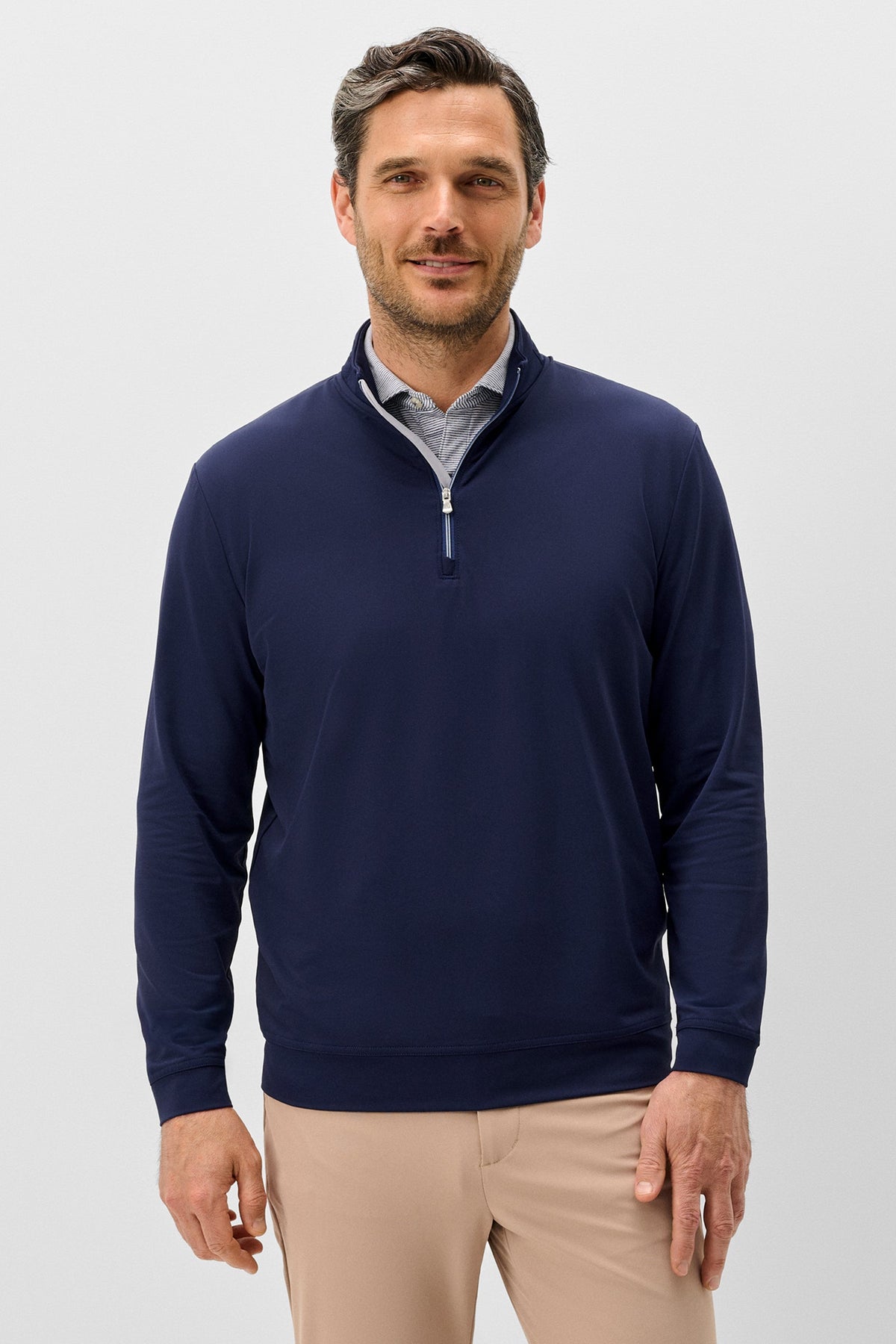 A man wearing a navy blue quarter-zip pullover sweater over a light blue patterned collared shirt and beige trousers, standing against a light gray studio background.