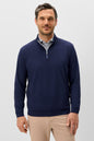 A man wearing a navy blue quarter-zip pullover sweater over a light blue patterned collared shirt and beige trousers, standing against a light gray studio background.