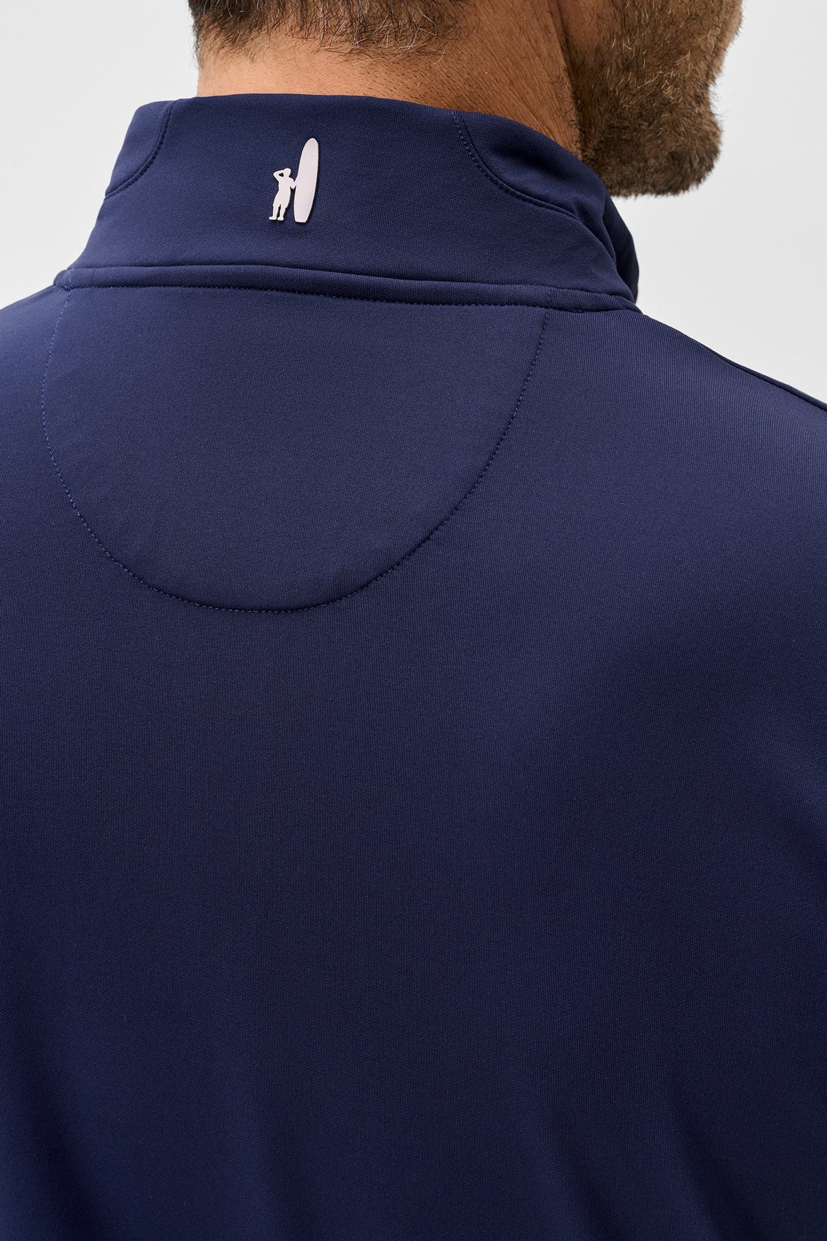 Close-up back view of a man wearing a navy blue high-neck athletic shirt with a small white logo on the collar, against a light gray background.