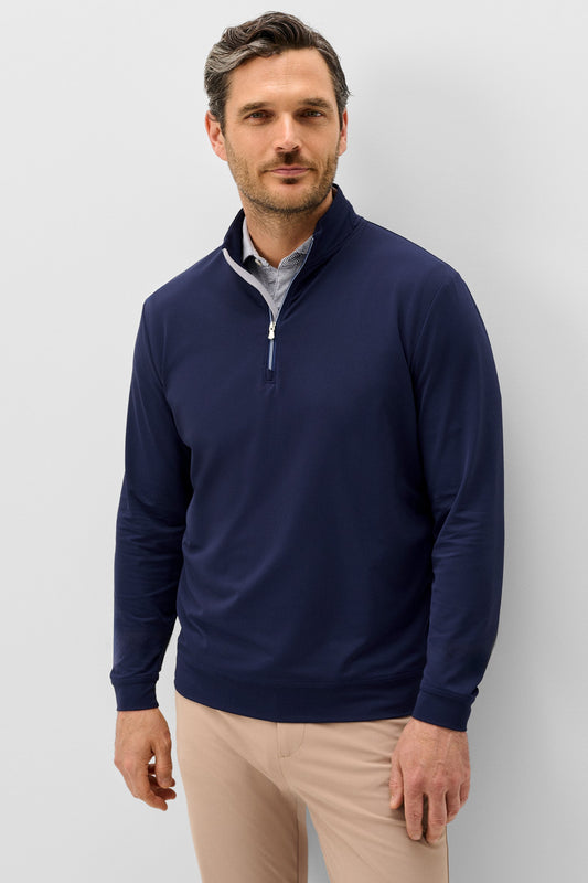 A man wearing a navy blue quarter-zip pullover sweater over a light blue collared shirt and beige trousers, standing against a light gray studio background.