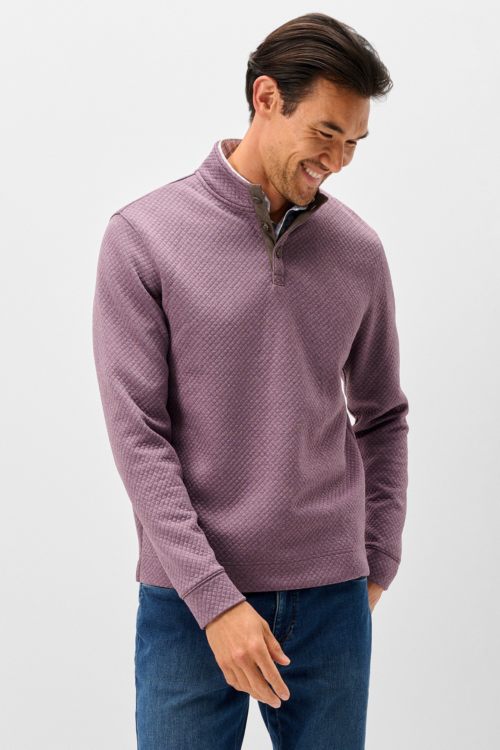 A man wearing a textured, waffle-knit quarter-zip pullover sweater in a dusty mauve-purple color, paired with blue jeans, photographed against a clean white studio background.