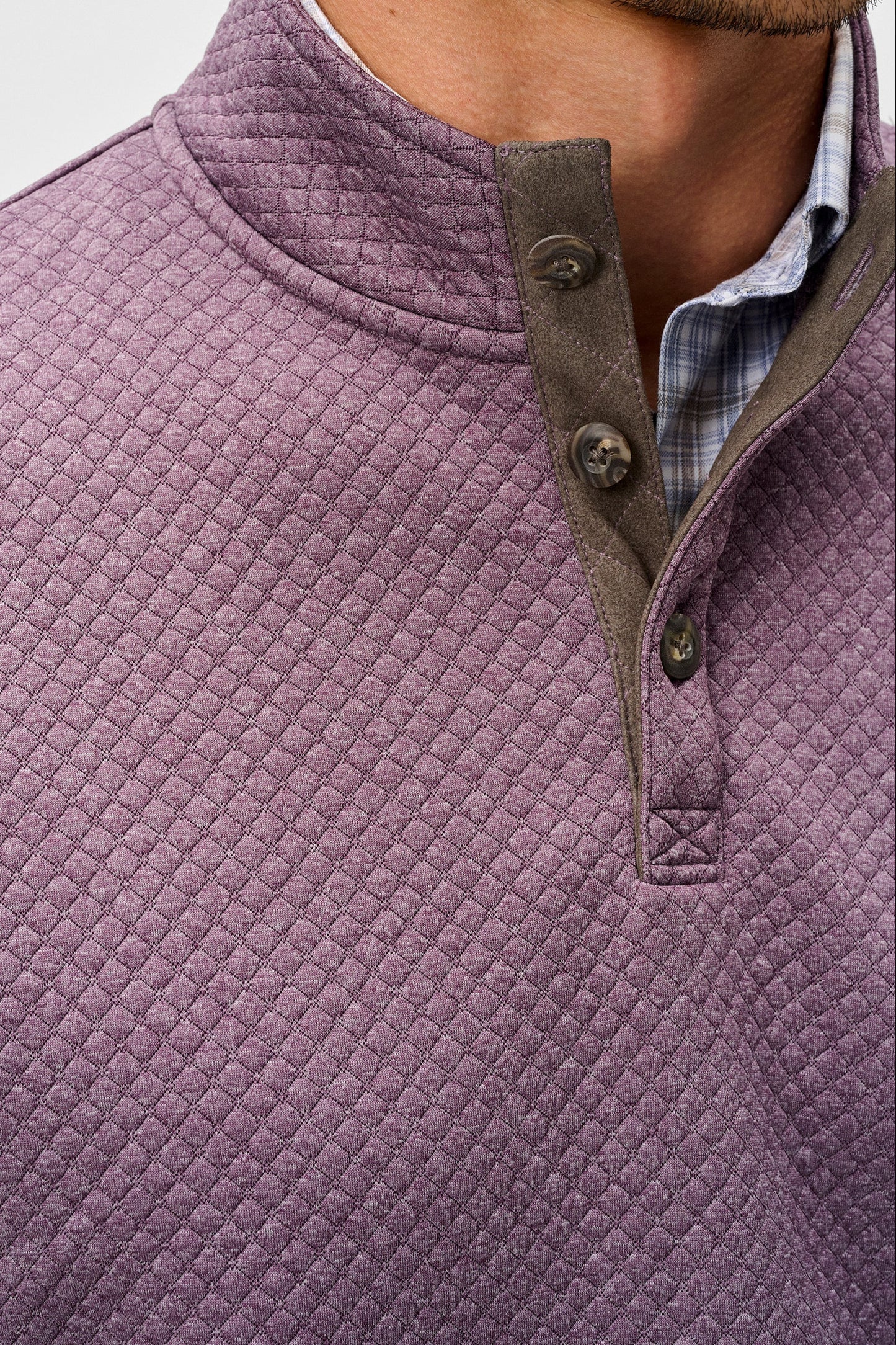 Close-up of a man wearing a textured, diamond-quilted pullover sweater in a dusty mauve-purple color, featuring a brown buttoned placket and collar trim over a blue plaid shirt.