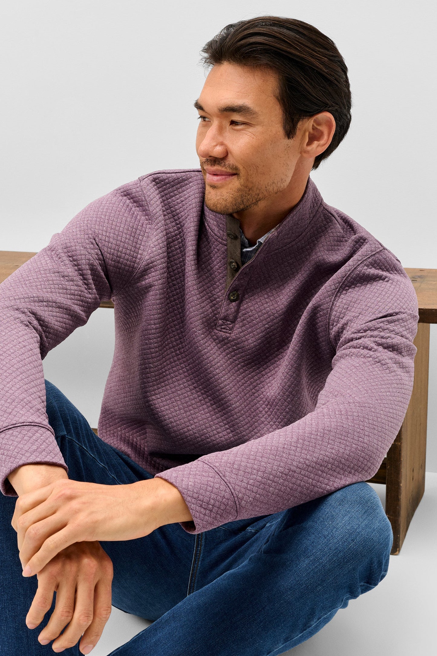 A man wearing a textured waffle-knit long-sleeve quarter-placket pullover in dusty purple, paired with blue jeans, sitting on a wooden bench against a light gray background.