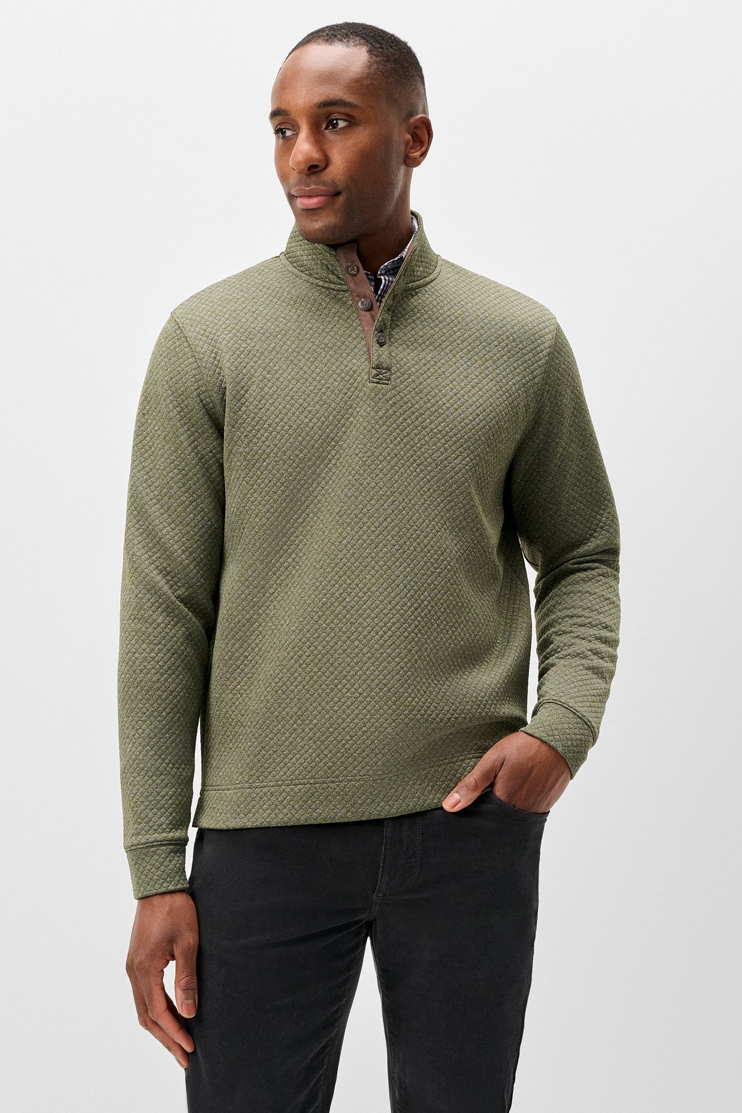A man wearing an olive green textured quarter-zip pullover sweater with a high collar, paired with black trousers, photographed against a light gray studio background.