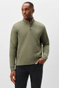 A man wearing an olive green textured quarter-zip pullover sweater with a high collar, paired with black trousers, photographed against a light gray studio background.