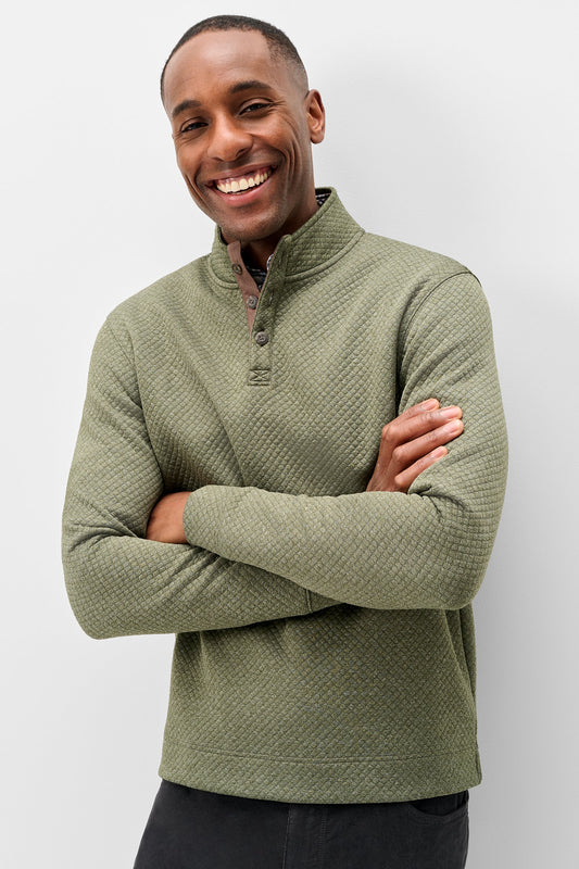 A smiling man wearing an olive green textured quarter-zip pullover sweater, standing with arms crossed against a light gray studio background.
