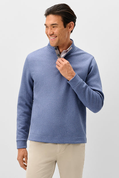 A man wearing a textured blue quarter-zip pullover sweater and beige pants, standing against a light gray studio background.