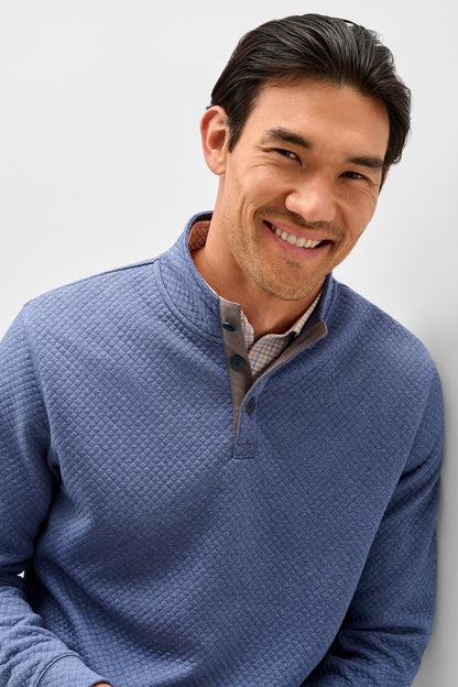 A smiling man wearing a textured blue quarter-zip pullover sweater over a collared shirt, photographed against a clean light gray background.