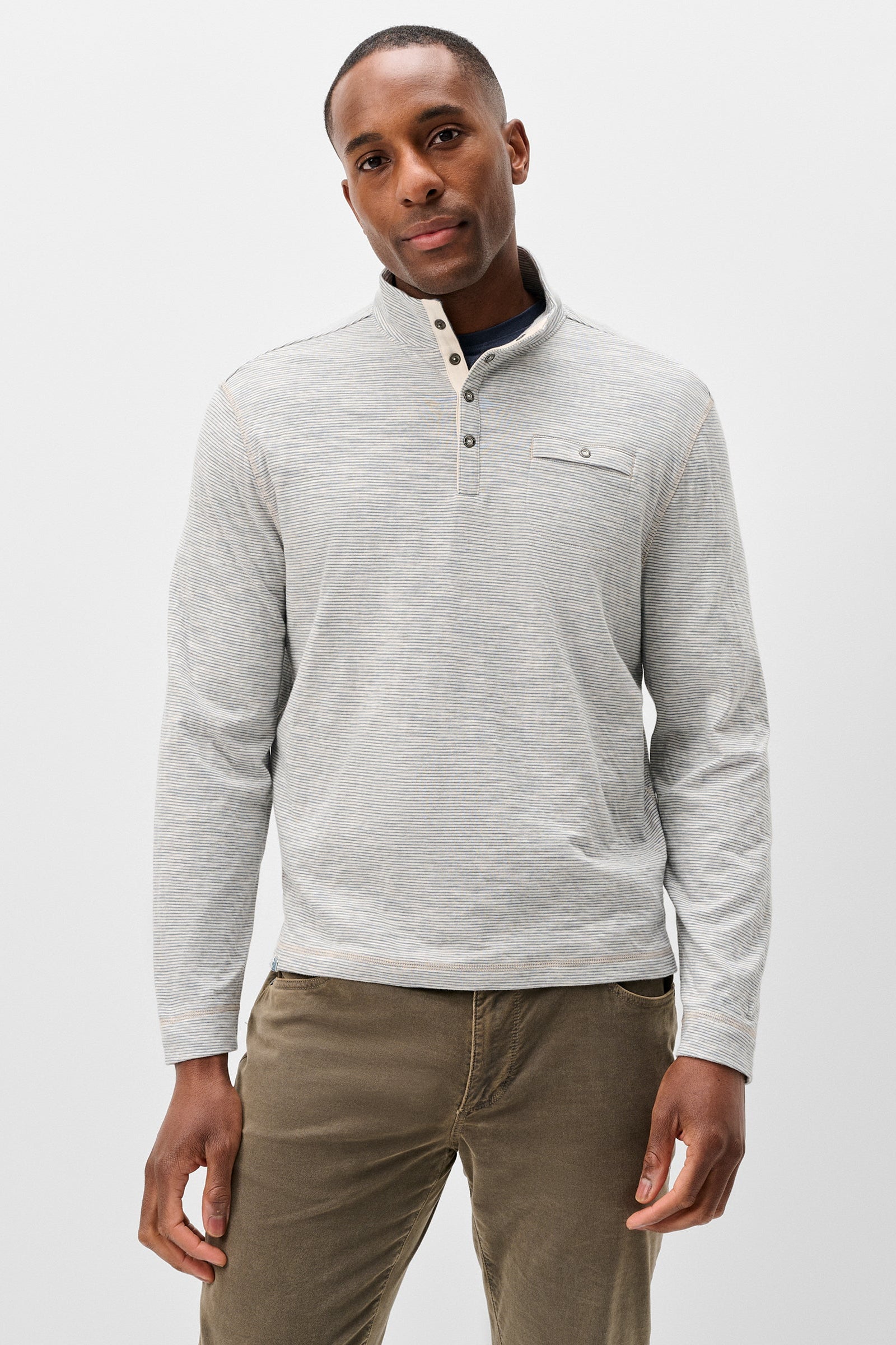 A man wearing a light gray heathered long-sleeve quarter-placket henley shirt with a small chest pocket, paired with brown pants, against a plain light gray studio background.