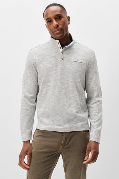 A man wearing a light gray heathered long-sleeve quarter-placket henley shirt with a small chest pocket, paired with brown pants, against a plain light gray studio background.