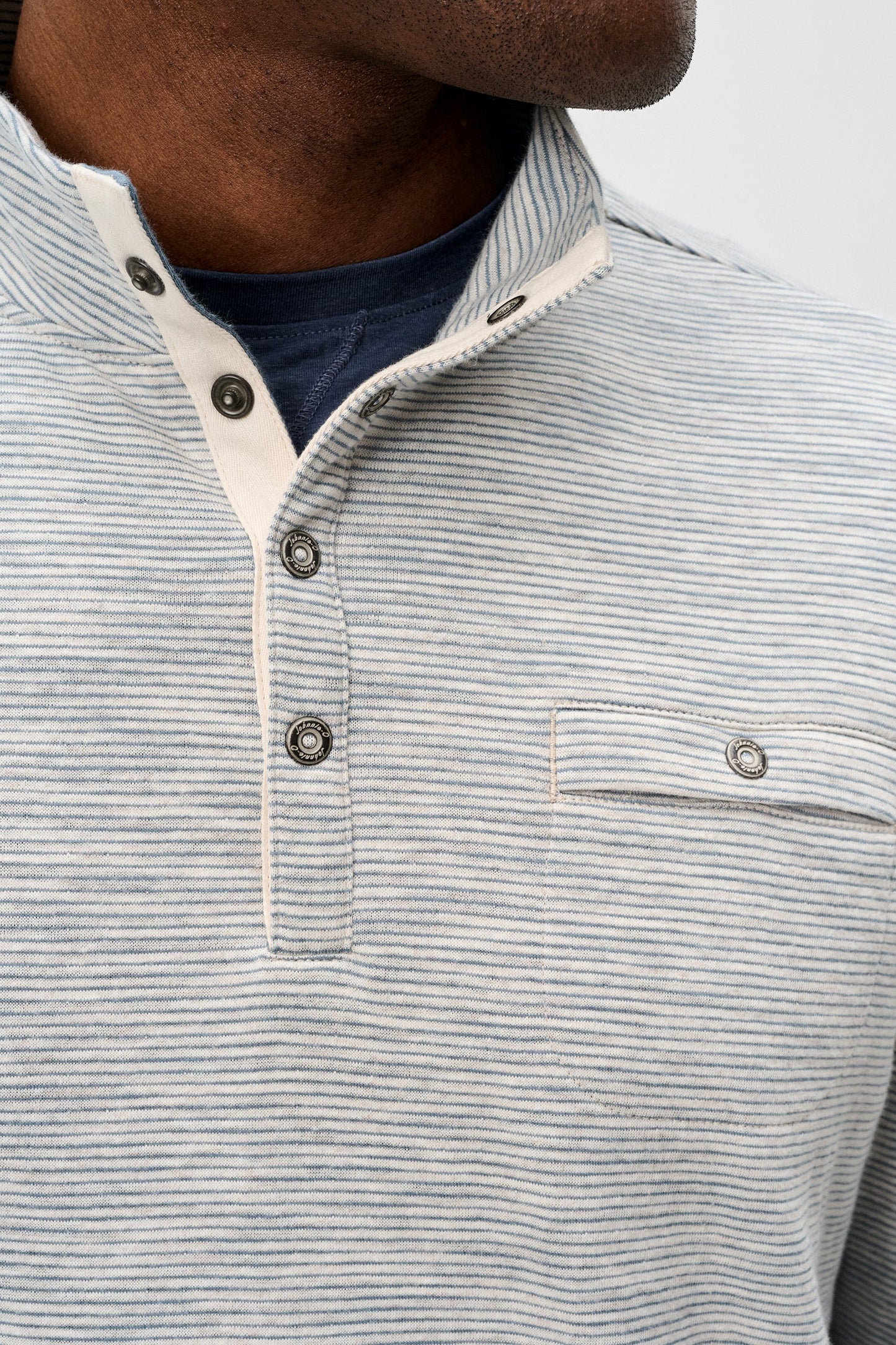 Close-up of a man wearing a light gray and white striped henley shirt with a buttoned collar and a chest pocket, layered over a navy blue t-shirt.
