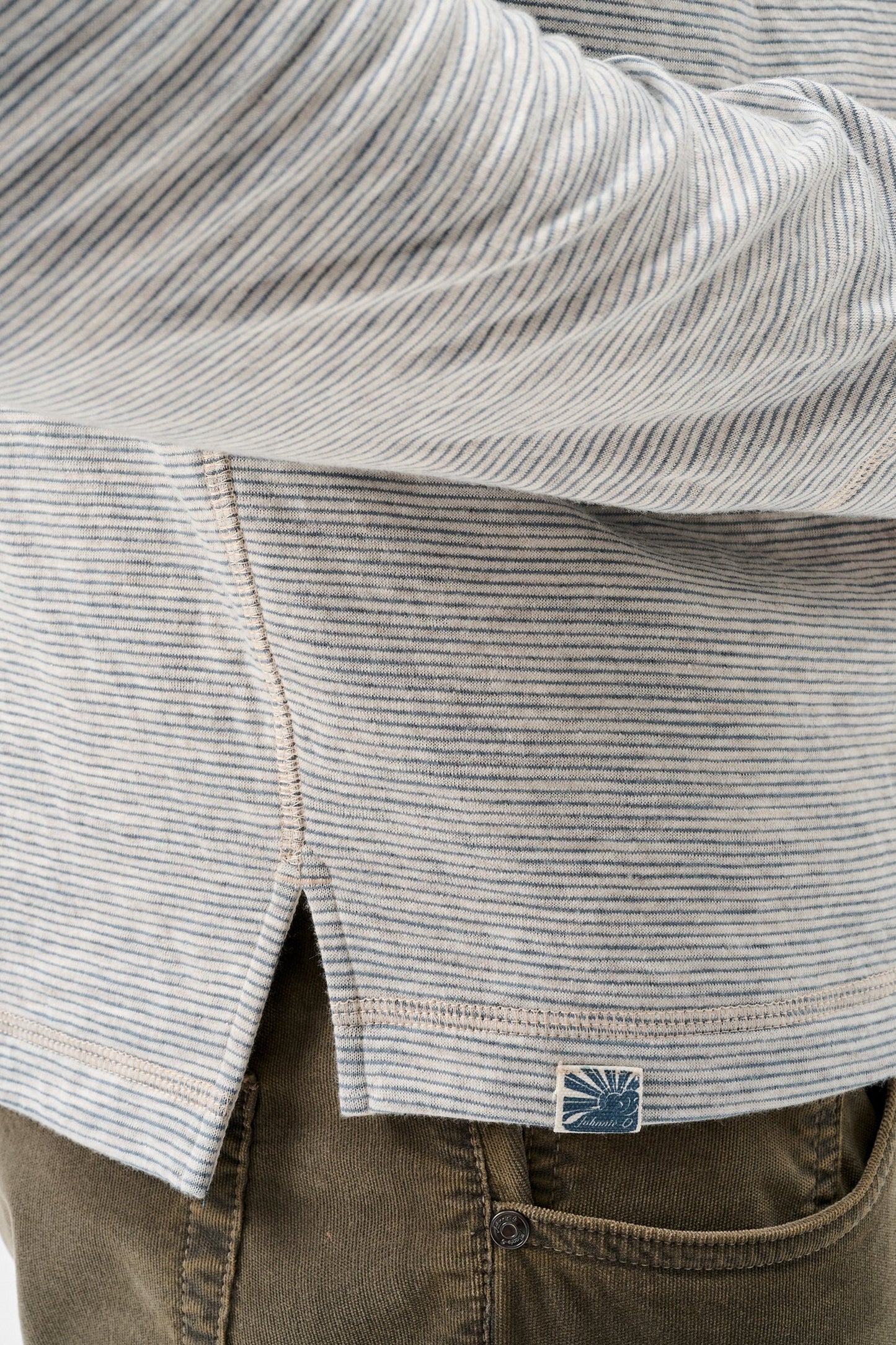 Close-up of a person wearing a light gray and white striped long-sleeve shirt with a small blue brand patch near the hem, paired with brown pants.