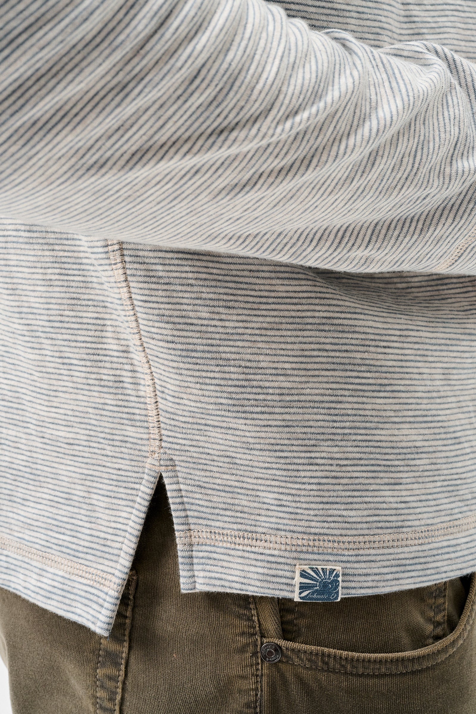Close-up of a person wearing a light gray and white striped long-sleeve shirt with a small blue brand patch near the hem, paired with brown pants.