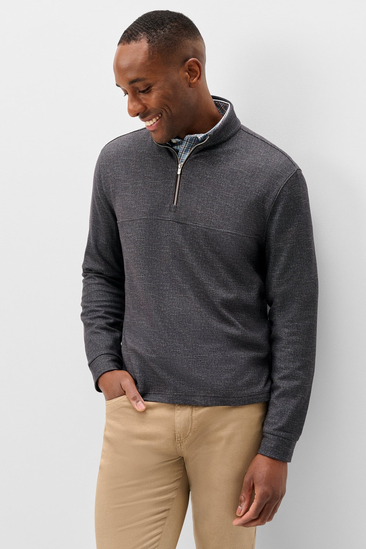 A man wearing a dark gray heathered quarter-zip pullover sweater over a light blue collared shirt, paired with khaki pants, standing against a light gray studio background.