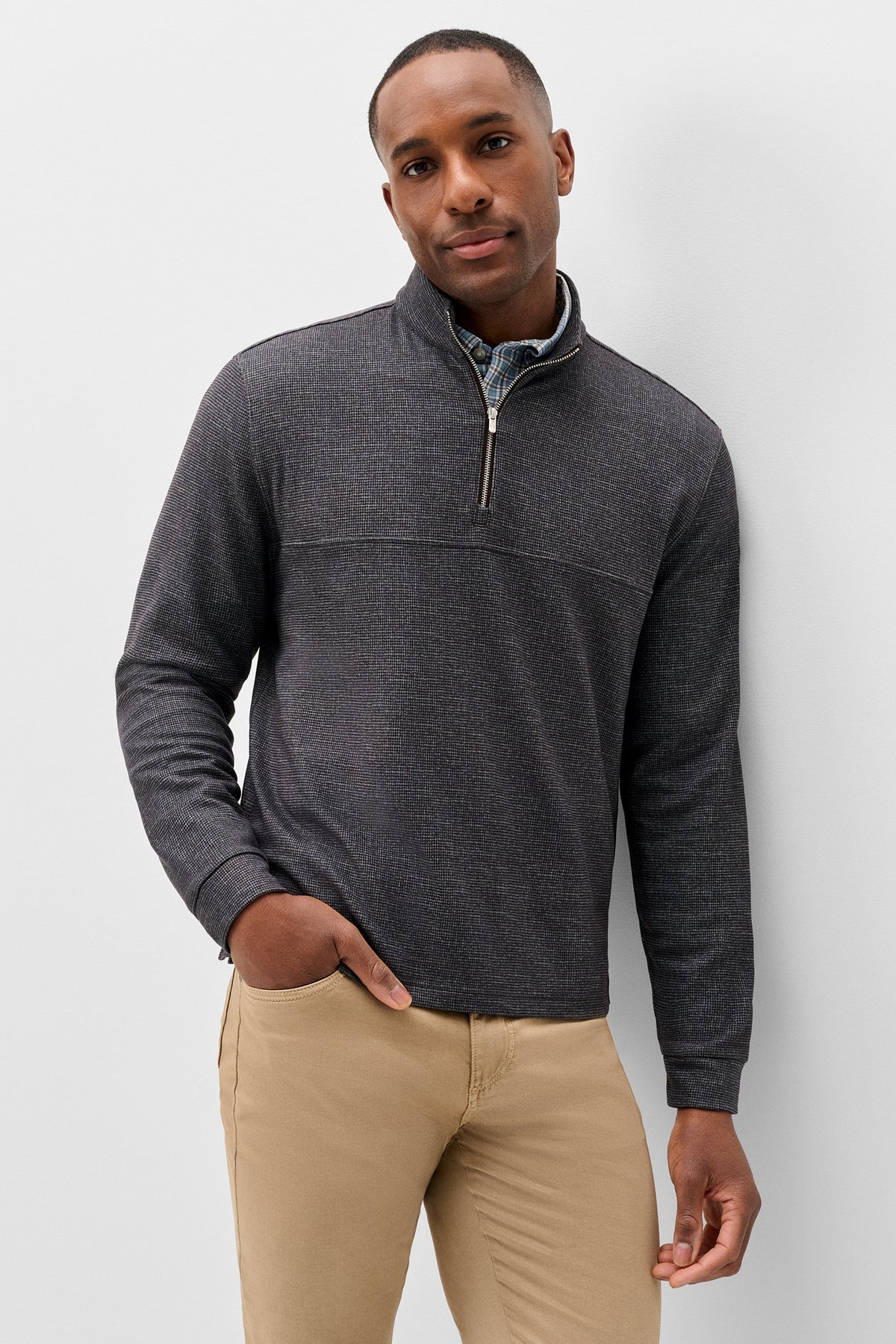 A man wearing a dark gray quarter-zip pullover sweater with a mock neck, paired with khaki pants, standing against a light gray studio background.
