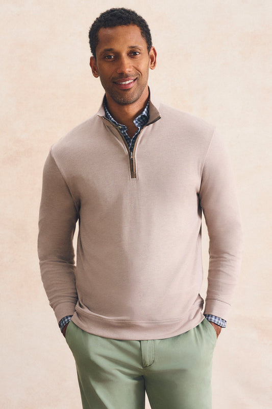 A man wearing a light beige quarter-zip sweater layered over a blue and white checkered collared shirt, paired with sage green trousers, standing against a textured beige background.