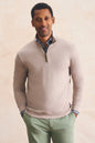 A man wearing a light beige quarter-zip sweater layered over a blue and white checkered collared shirt, paired with sage green trousers, standing against a textured beige background.