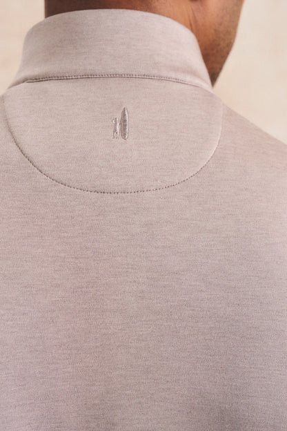 Close-up back view of a person wearing a light taupe heathered knit turtleneck sweater, showing the collar and a small embroidered logo.