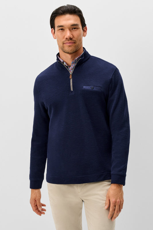 A man wearing a navy blue quarter-zip pullover sweater with a small chest pocket, layered over a patterned collared shirt and paired with beige trousers, photographed against a light gray studio background.