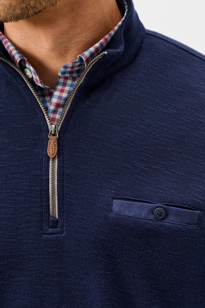 Close-up of a man wearing a navy blue quarter-zip pullover sweater over a red, white, and blue plaid collared shirt. The sweater features a ribbed texture, a buttoned chest pocket, and a brown leather pull tab on the zipper.