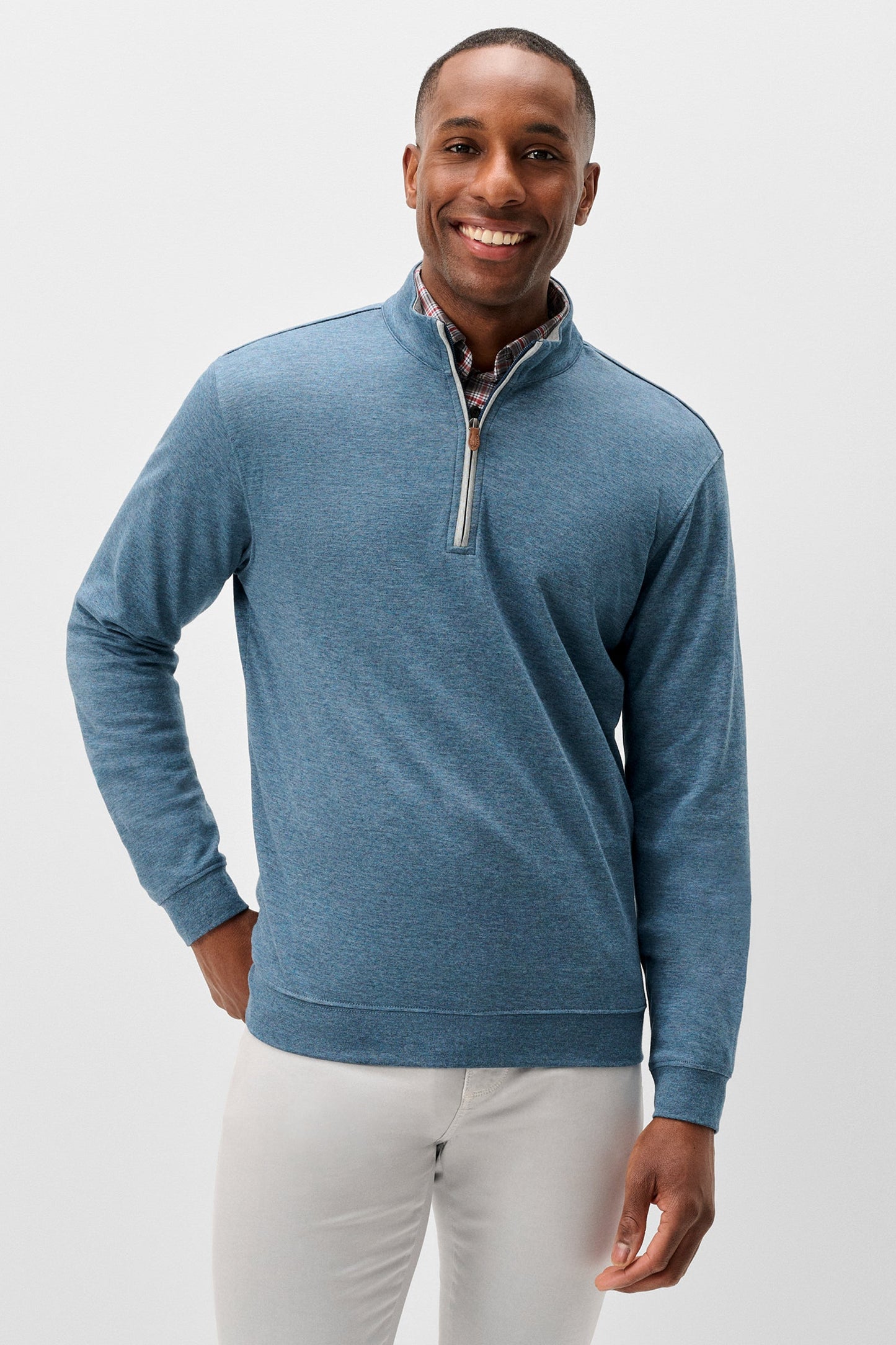 A smiling man wearing a heathered blue quarter-zip pullover sweater with a patterned inner collar and white trousers, standing against a light gray studio background.