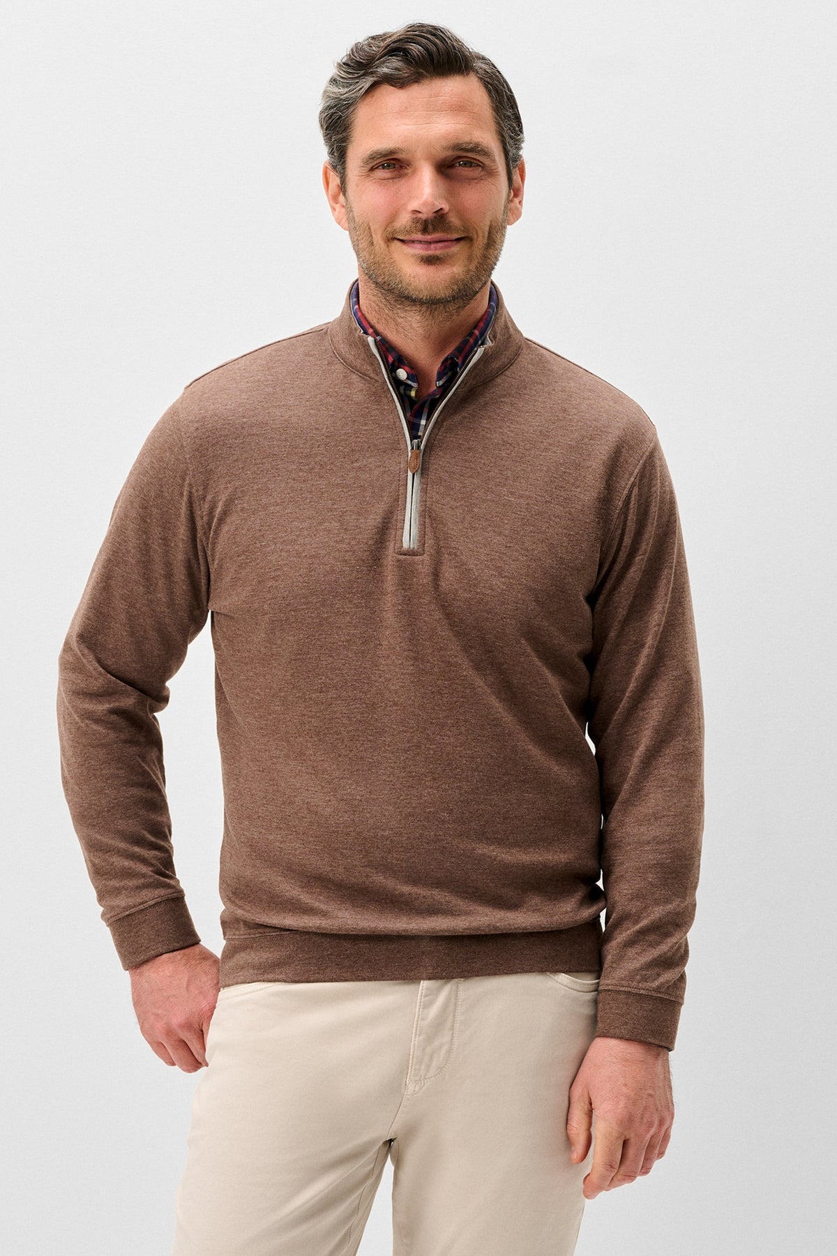 A man wearing a brown quarter-zip knit sweater over a patterned collared shirt and beige trousers, photographed against a light gray studio background.