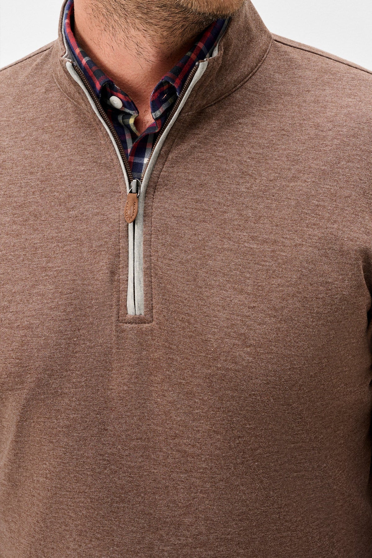 A close-up of a man wearing a heathered brown quarter-zip pullover sweater, layered over a red and blue plaid collared shirt. The sweater features a stand collar, a silver zipper, and a small brown leather pull tab.